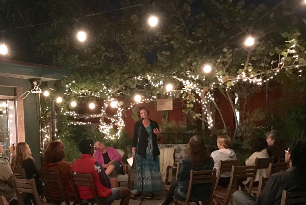 Stephanie standing performing poetry to people sitting in chairs with a background of trees with white lights draped across