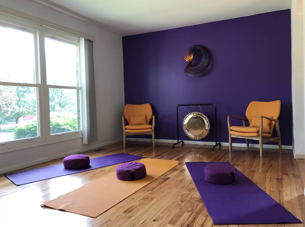 Studio space with hardwood floors, purple wall, a spiral sculpture on the wall, orange chairs, a gong and yoga mats with cushions