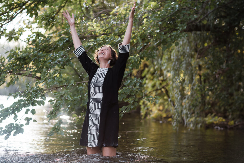 queer white cis woman with arms to sky throwing water wearing a short dress with legs in the Huron River