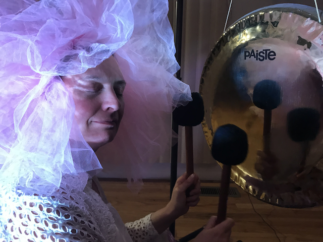 Events Stephanie with pink tutu around face, closed eyes, playing a gold reflective gong with balck mallets