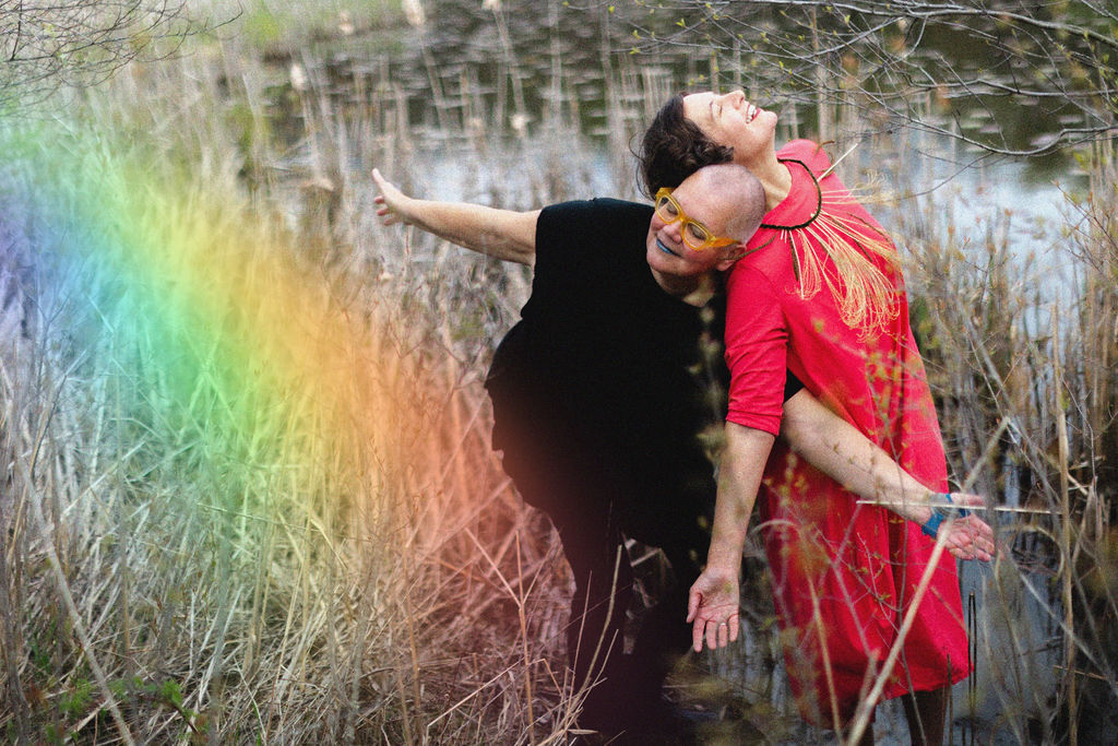Petra in black and Stephanie in coral, leaning into each other in interdependent delight amidst early spring grasses at LeFurge Preserve in Ypsilanti with rainbow magic overlay. 