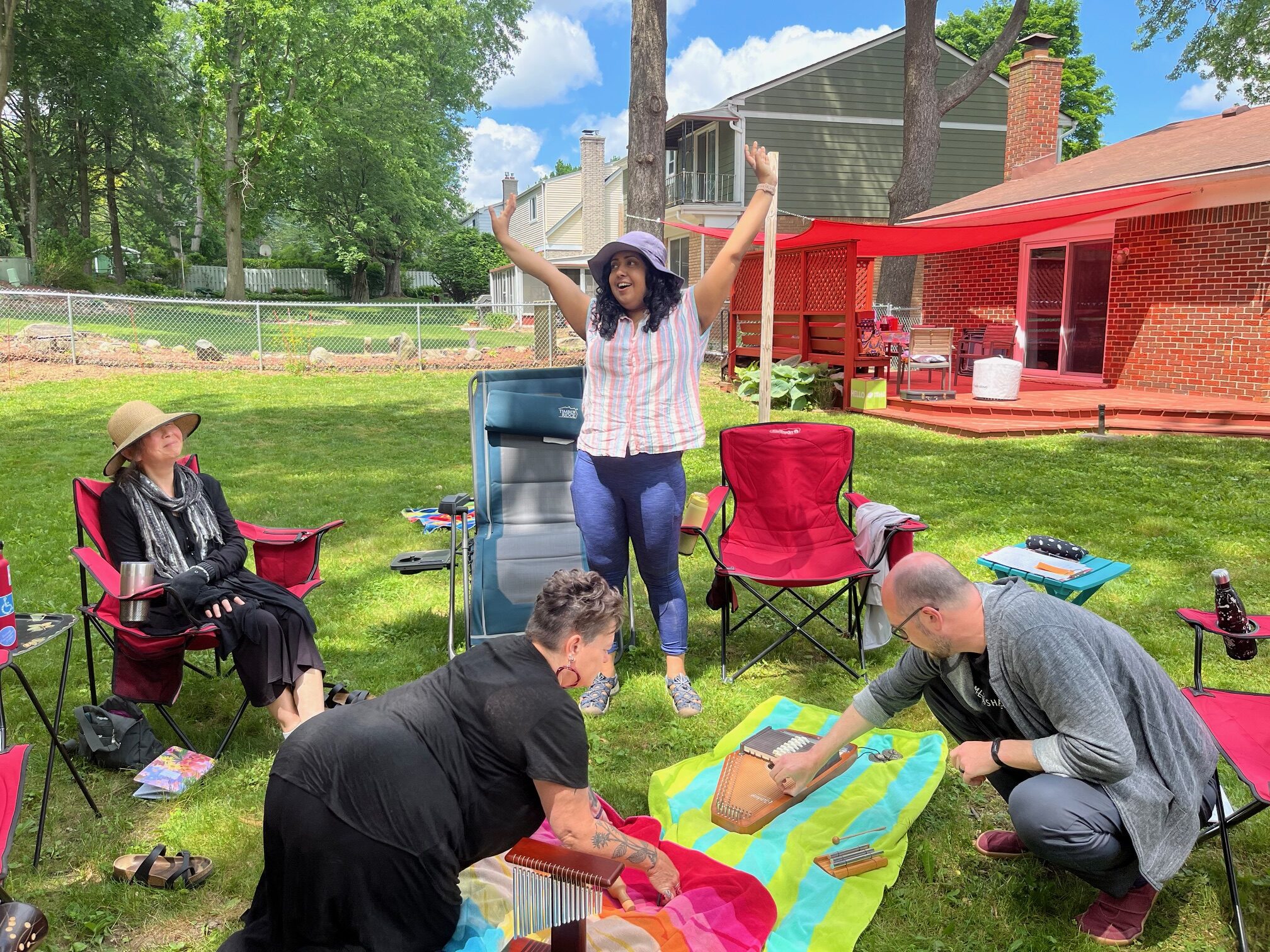 Four people in a backyard, one has her arms up reaching to the sky and  smiling, another is sitting on a red camping chair humming, and the other two people are on the ground reaching toward various instruments. 