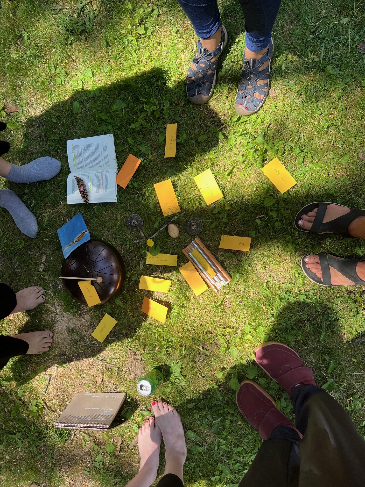 Outdoors on grass with sun and shadows, a circle of six pairs of feet encircling a mix of yellow paper scraps with words, a steel drum, notebook, pine cone, Le Croix can 